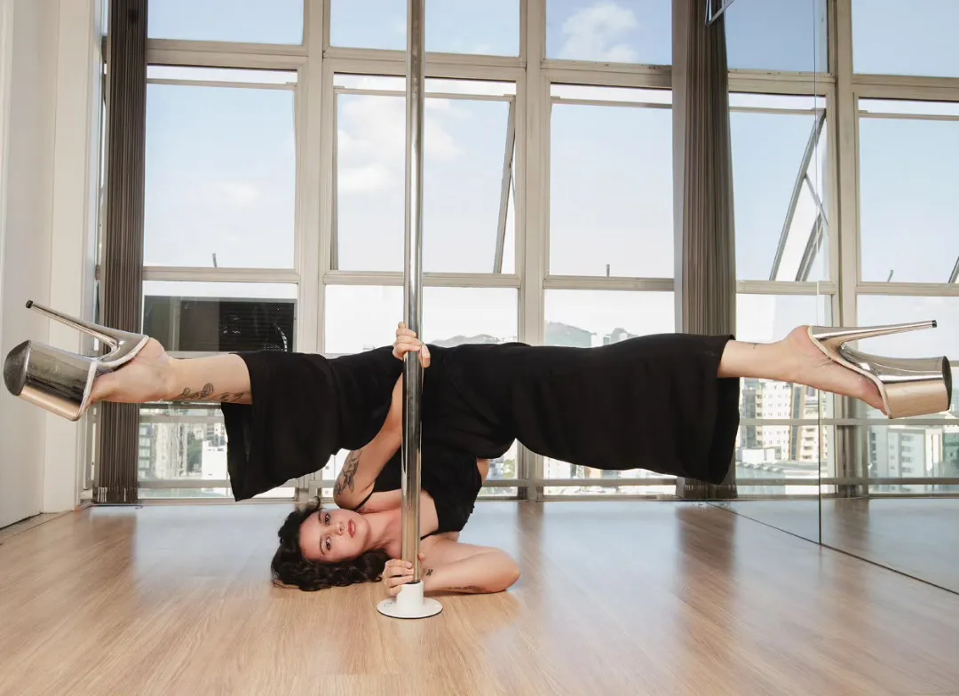 Aluna deitada no chão com as pernas abertas de salto alto ao redor do pole, no estúdio Old School Pole Dance BH, com janelas amplas ao fundo